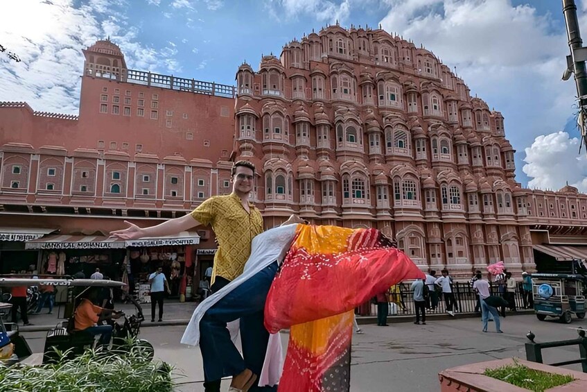 A person in a yellow shirt and white scarf standing in front of a large pink palace with multiple windows and balconies. 3