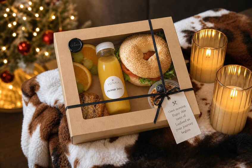 A gift box with a bagel, orange juice, and a candle, placed on a fur blanket with a Christmas tree in the background. 3
