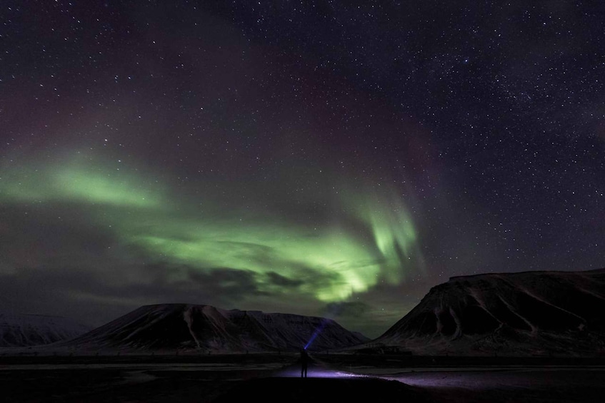 A person standing by a reflective water surface under a night sky with visible stars and the aurora borealis. 1