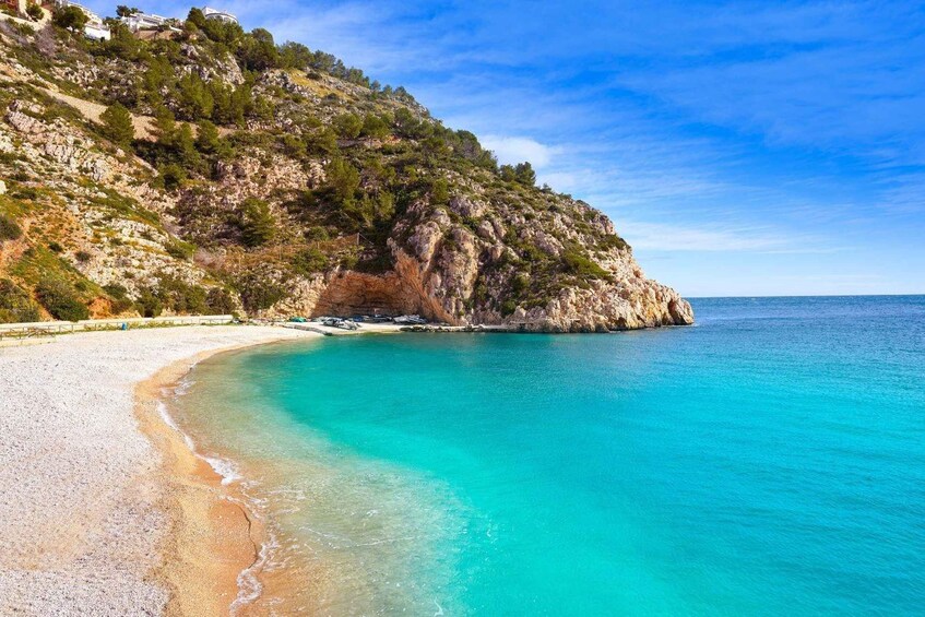 A coastal beach with clear turquoise waters, a sandy shore, and a rocky hillside with sparse vegetation. 2