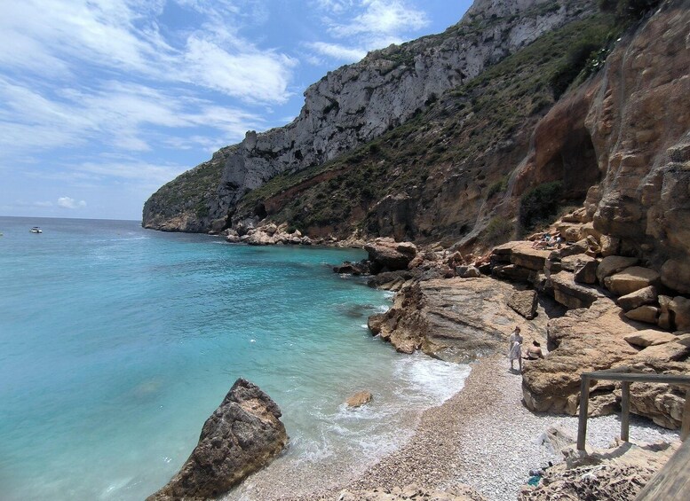 A rocky coastline with clear turquoise waters, a small boat, and a person walking along the shore. 1