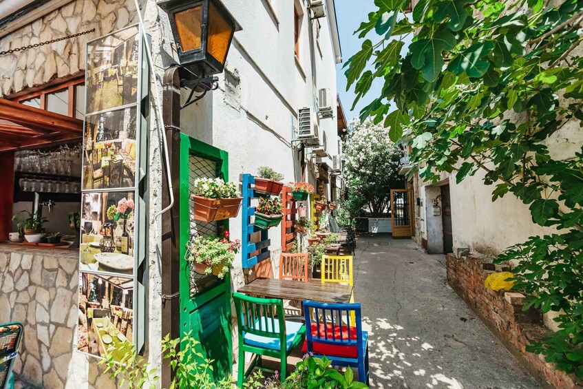 A narrow street with colorful outdoor seating, potted plants, and a stone building facade. 5