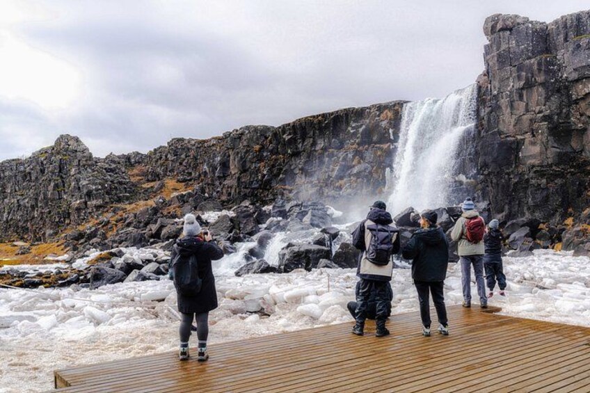 A group of people standing on a wooden platform, observing a waterfall cascading down a rocky cliff. 4