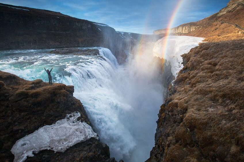 A waterfall with a person standing on a rock, a rainbow, and a rugged landscape. 1