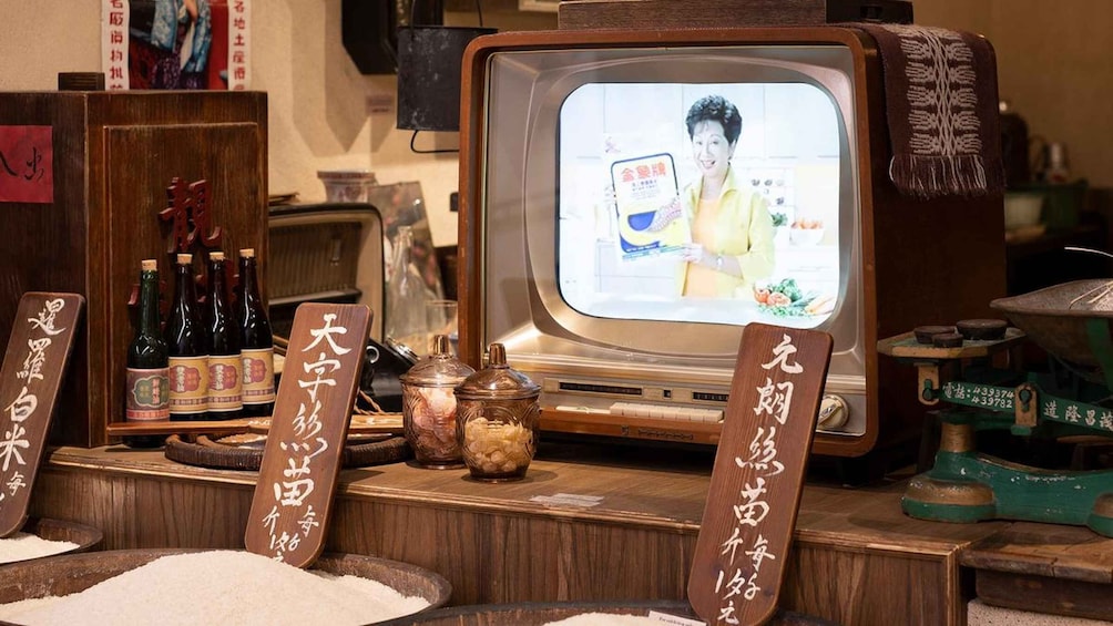 A vintage television set displaying a person, surrounded by traditional Japanese items like sake bottles and wooden signs with Japanese characters. 4