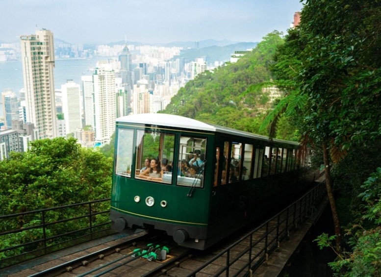 A green cable car with passengers, traveling along a track surrounded by greenery and overlooking a cityscape. 1