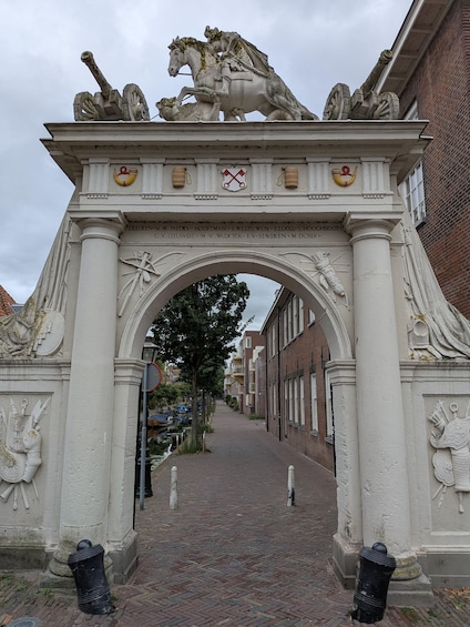 An ornate stone archway with sculptures of horses and riders, flanked by columns with inscriptions, leading into a brick-paved street. 3