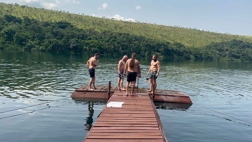 A group of people standing on a wooden dock over calm water with a forested hill in the background. 3