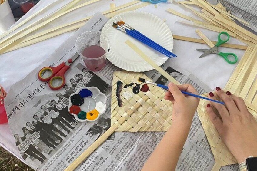 A person weaving with bamboo strips on a newspaper-covered surface, surrounded by scissors, paint, and a plate. 3