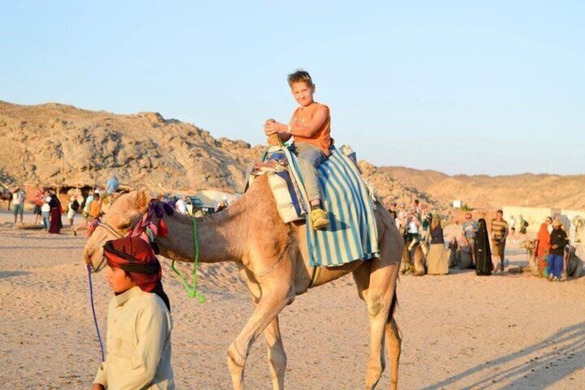 A camel with a rider in a desert setting, surrounded by people. 4