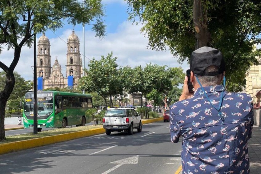 Un autobús verde, un coche blanco y una persona tomando una fotografía en una calle de la ciudad con un edificio histórico al fondo. 3