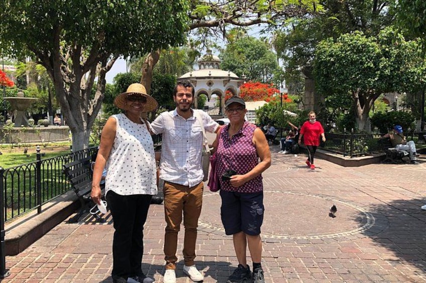 Trois personnes se tiennent sur un chemin pavé dans un parc, avec des arbres et une fontaine en arrière-plan. 4