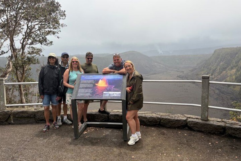 A group of people standing by a rocky overlook with a large informational sign and a view of a volcanic landscape. 2