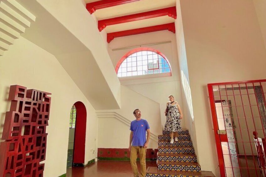 Un hombre y una mujer en una escalera dentro de un edificio con paredes rojas y blancas, un amplio ventanal de arco y un adorno mural geométrico. 1