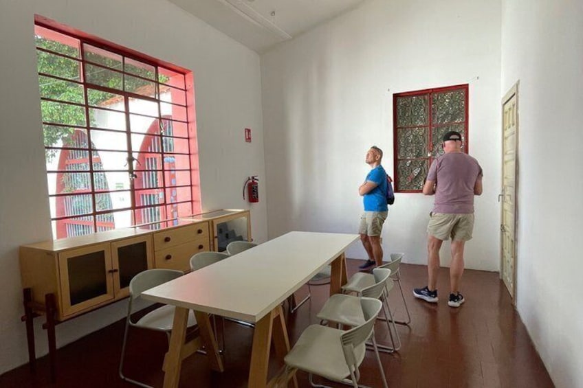 Une salle de classe avec une longue table, des chaises, une fenêtre aux montants rouges et une porte. 4