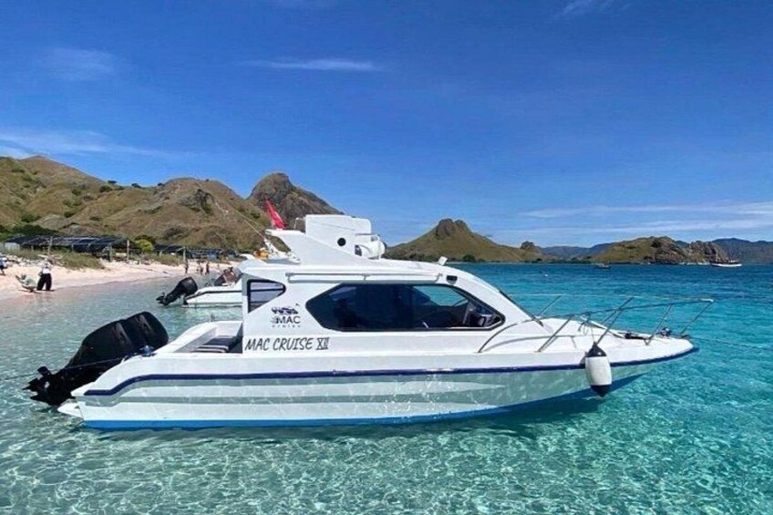 En vit båt med namnet ”MAC CRUISE XII” lade till vid en strand med kristallklart turkosfärgat vatten och avlägsna kullar i bakgrunden. 5