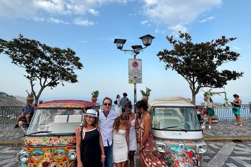 Un gruppo di persone in piedi tra due auto d'epoca colorate, parcheggiate su una strada acciottolata, con alberi e un lampione sullo sfondo. 1