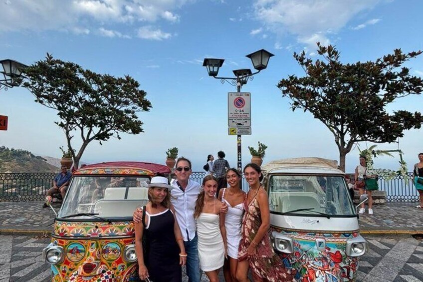 Un gruppo di persone che posa davanti a due auto d'epoca colorate su una superficie pavimentata con alberi e cielo sereno. 2