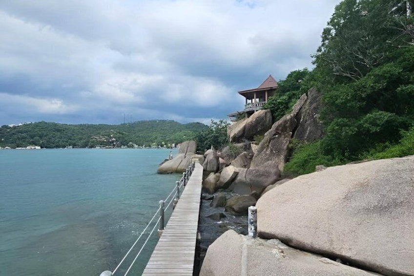 Eine Holzstege ragt in ruhiges Wasser hinein, große Felsen und ein Gebäude auf einem Hügel. 2