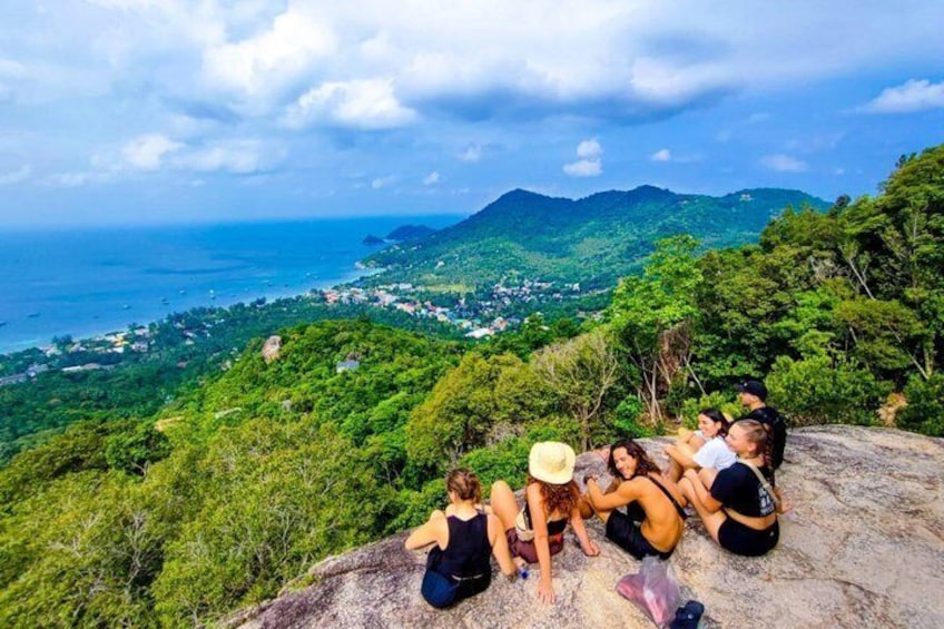 Eine Gruppe von Menschen sitzt auf einem Felsen und blickt auf eine Küstenlandschaft mit Bergen und viel Grün. 1