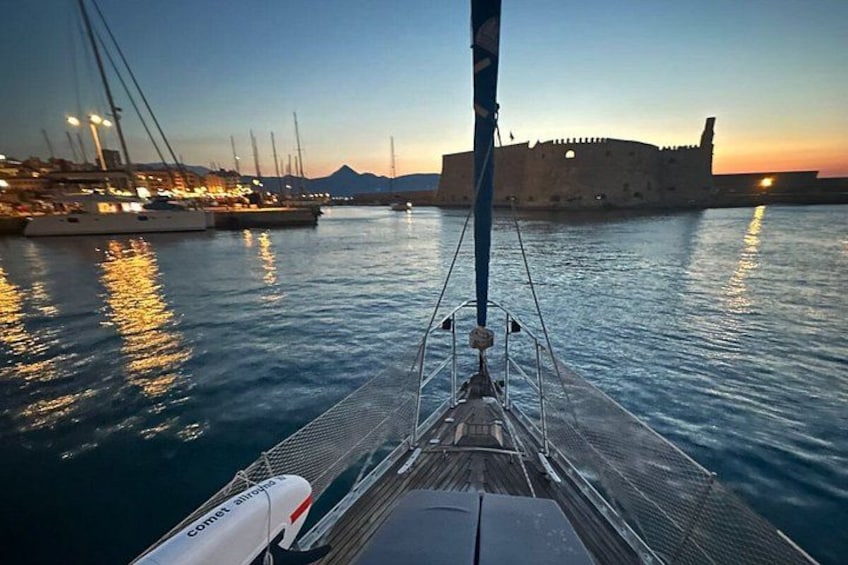 Cubierta de un velero con vista a un puerto al atardecer, un castillo en el fondo y barcos amarrados a lo largo de la orilla. 2