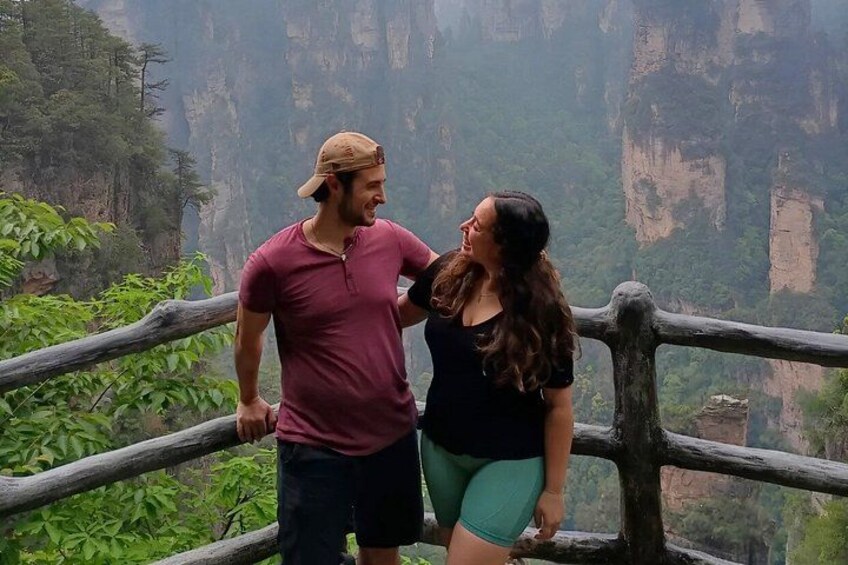 A couple standing on a wooden railing overlooking a canyon with steep cliffs and dense greenery. 5