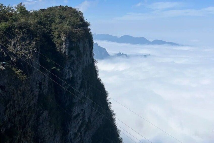 A mountain cliff with power lines, a forested area, and a layer of clouds below. 4