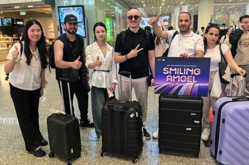 A group of people with luggage at an airport, holding a sign that reads 'SMILING ANGEL.' 2
