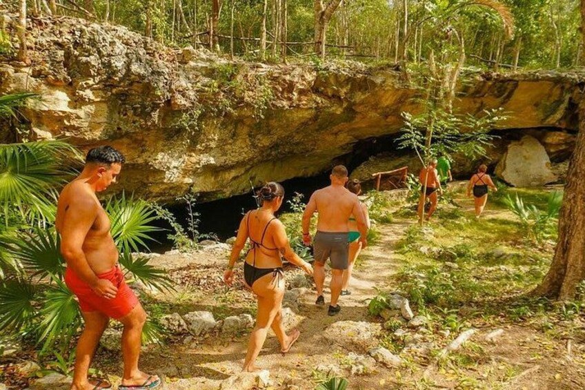 Eine Gruppe von Menschen geht auf einem steinigen Pfad entlang, umgeben von viel Grün und einem Höhleneingang. 2