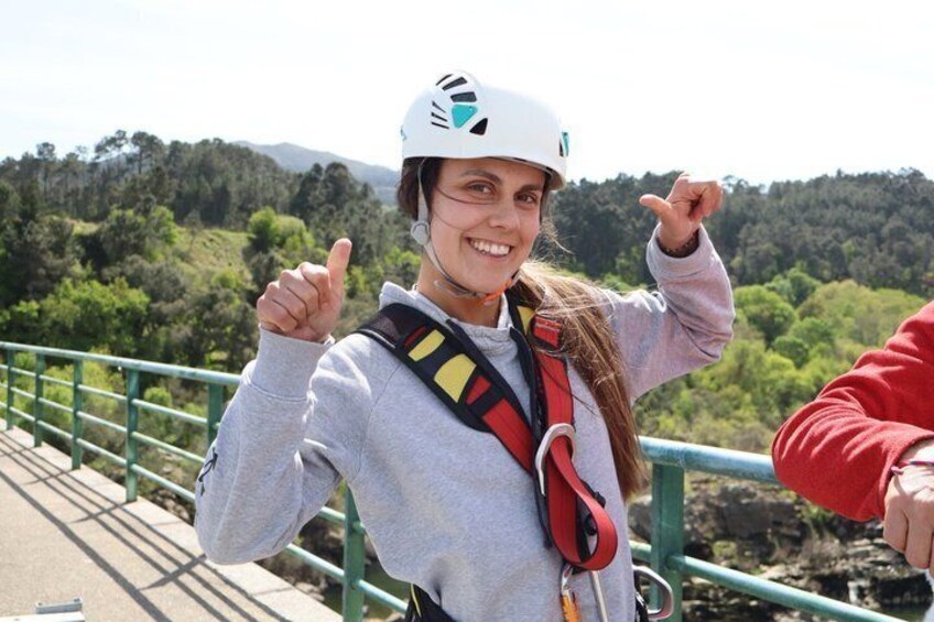 A person wearing a helmet and harness, standing on a bridge with greenery in the background. 4