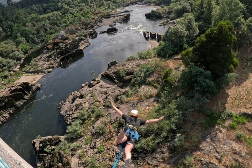 A person bungee jumping from a bridge over a river with surrounding greenery. 1