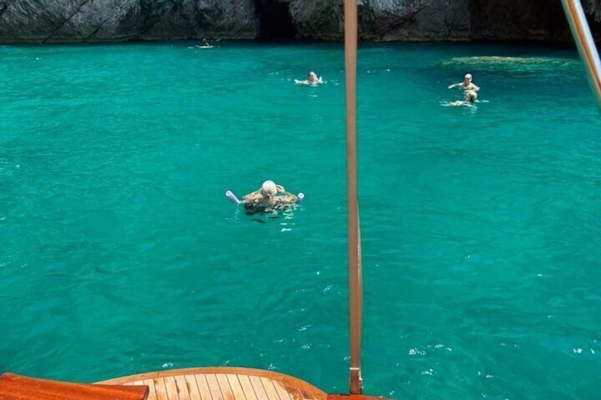 A person swimming in clear turquoise water, a wooden boat deck, and rocky cliffs in the background. 3