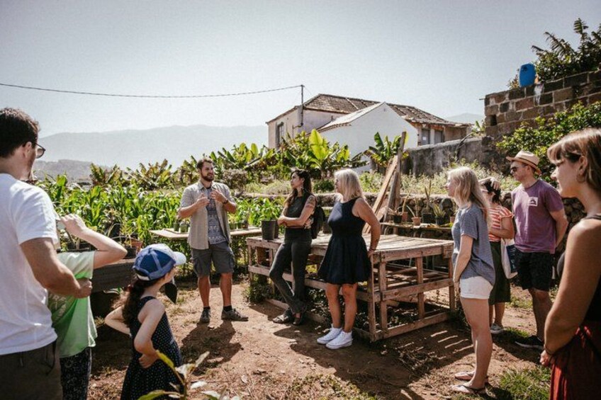 Un grupo de personas reunidas al aire libre, algunas de pie y otras sentadas, con una estructura de madera y plantas al fondo. 1