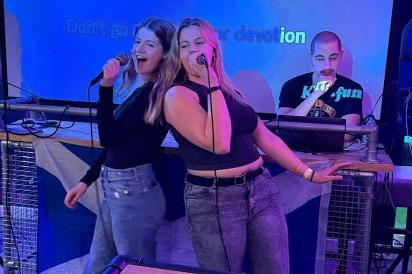 Two people singing on stage with a DJ in the background. 1