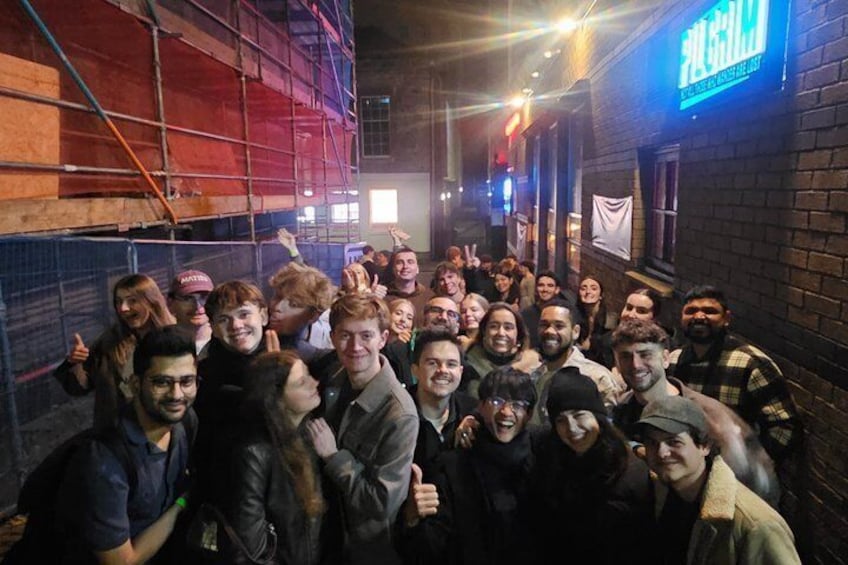 A group of people posing in an urban alleyway with brick walls and a neon sign. 5