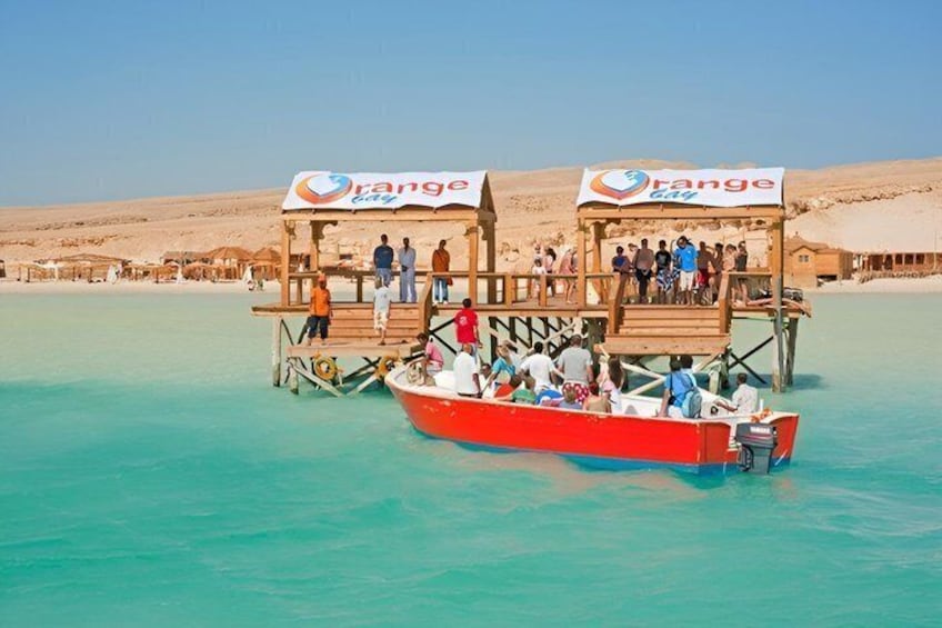Ein rotes Boot mit einer Holzkonstruktion auf einem türkisblauen Meer, ein Sandstrand und eine Wüste im Hintergrund. 4