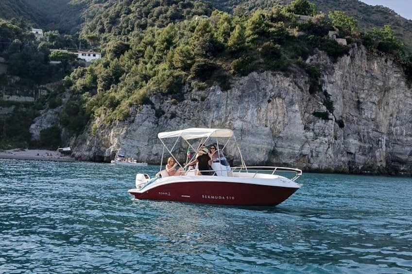 A red boat with 'KEMUDA STO' on the side, sailing on calm water near a rocky coastline with greenery and buildings in the background. 3