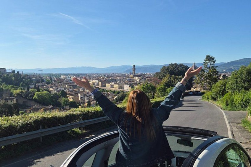 A person in a convertible car with arms outstretched, overlooking a cityscape with mountains in the background. 1