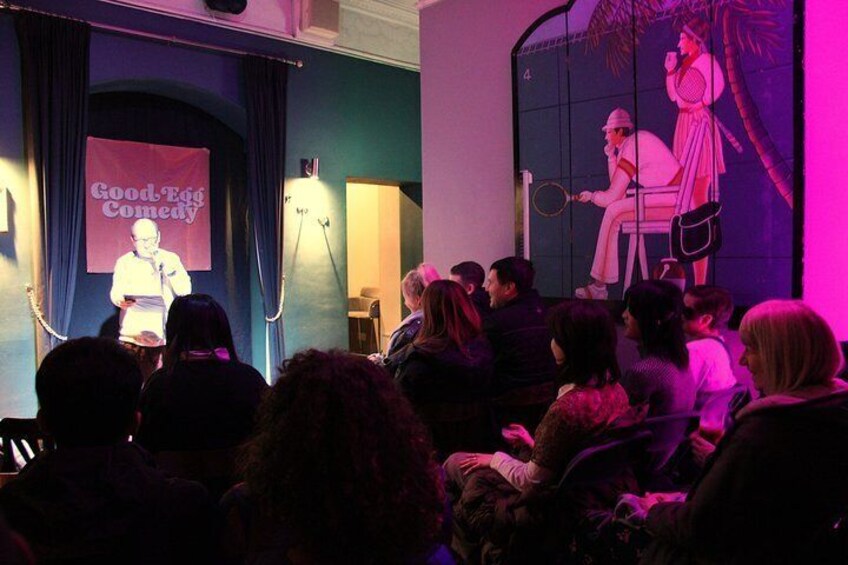 A stage with a banner reading 'Good Egg Comedy' and a person speaking to an audience. 1