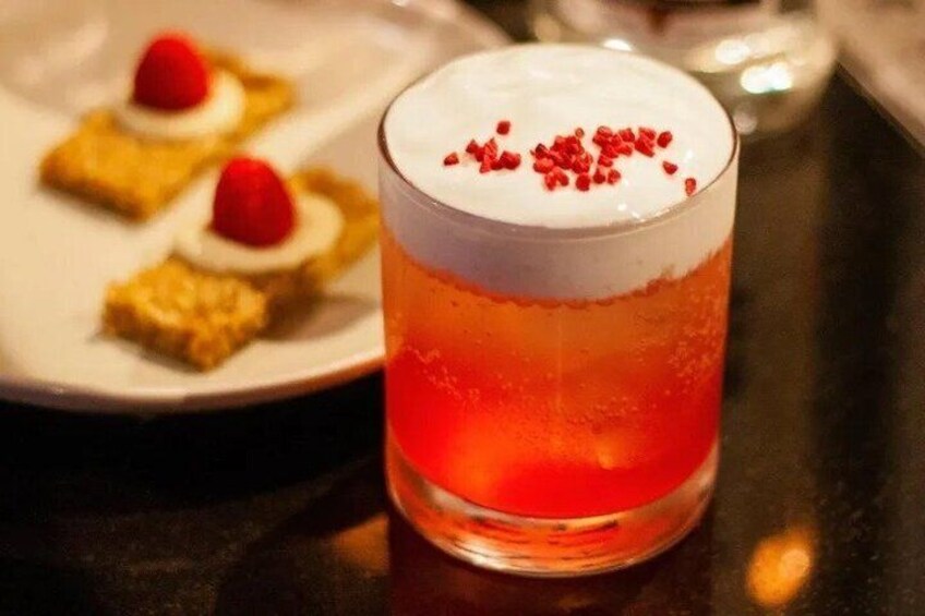A cocktail with a layered orange liquid, topped with white foam and red sprinkles, and a plate with small desserts in the background. 2
