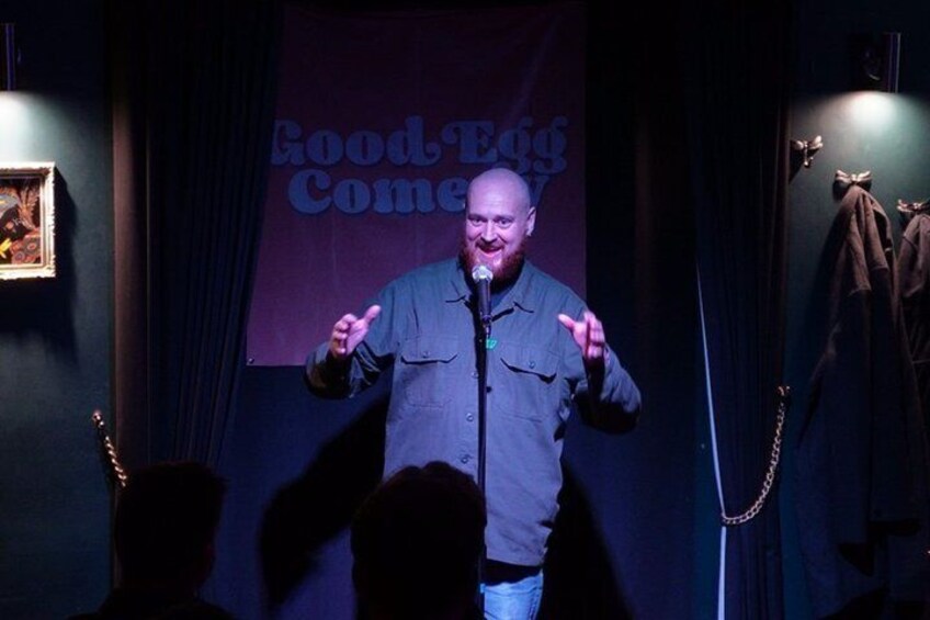 A person on stage with a microphone, a banner reading 'Good Egg Comedy,' and a dark background. 3