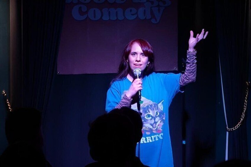 A person on stage with a microphone, wearing a blue shirt with a cat design, and a banner in the background that says 'Comedy'. 5