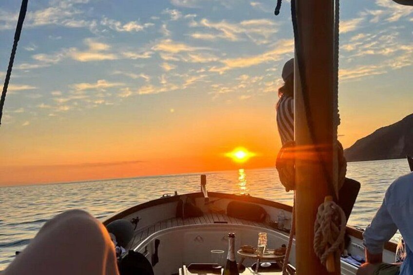 En båd i solnedgang med en person stående på dækket, et reb hængende fra masten og roligt vand. 5