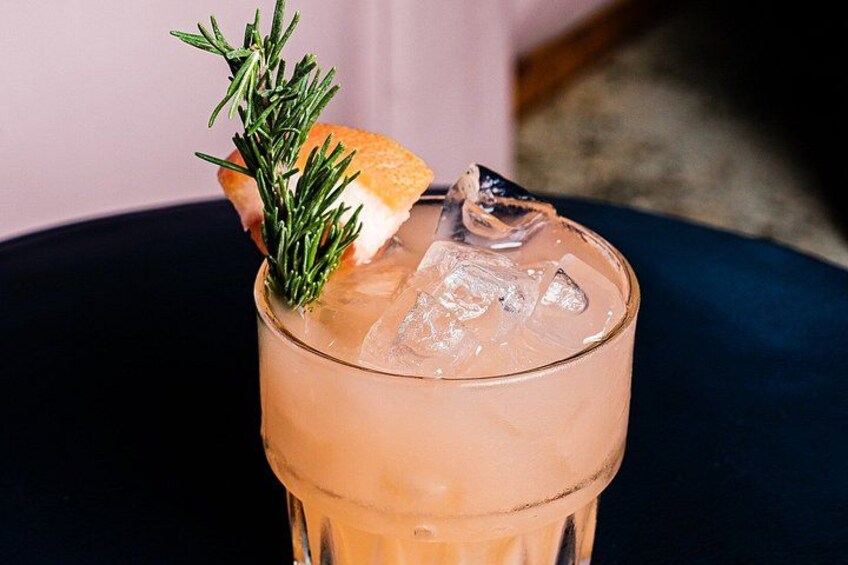 A glass of a pinkish beverage with ice cubes, garnished with a sprig of rosemary and a slice of orange. 2