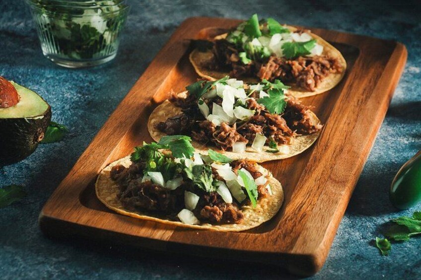 Three tacos on a wooden board with shredded meat, diced onions, and cilantro. 2