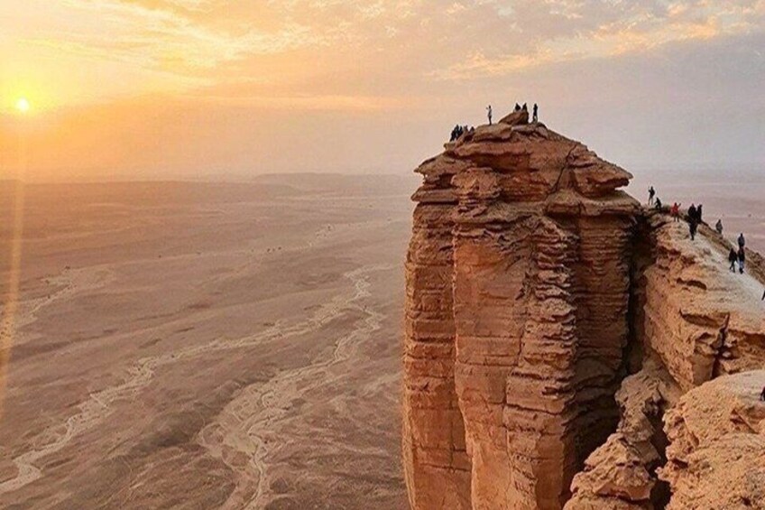 A rock formation with people at the top, a vast desert landscape, and the sun setting in the background. 1