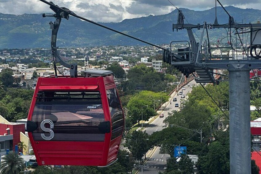 Un téléphérique rouge suspendu à des câbles, une route avec des véhicules, et un paysage urbain avec des bâtiments et des montagnes en arrière-plan. 4