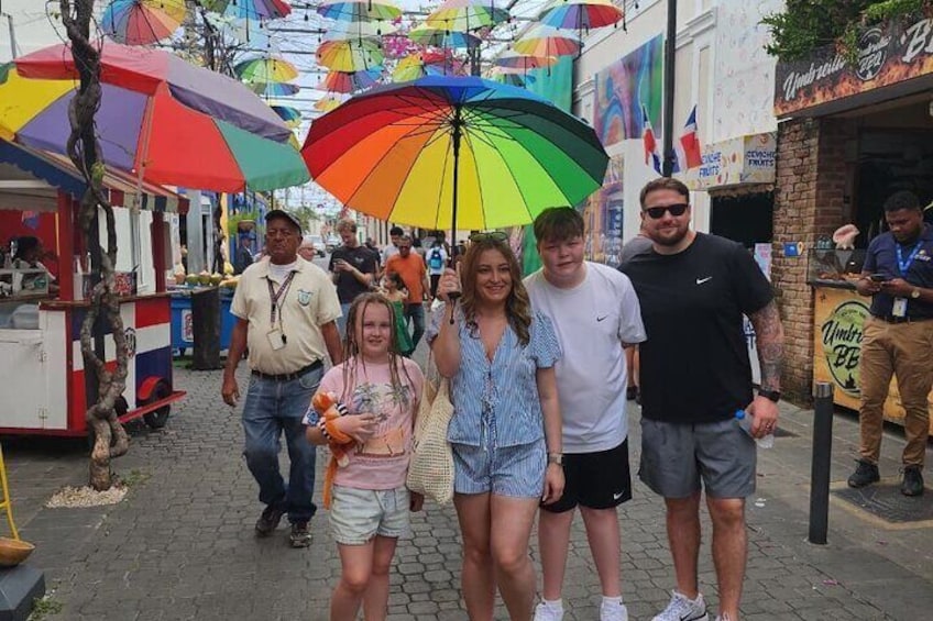 A street market with colorful umbrellas, food stalls, and people walking. 1