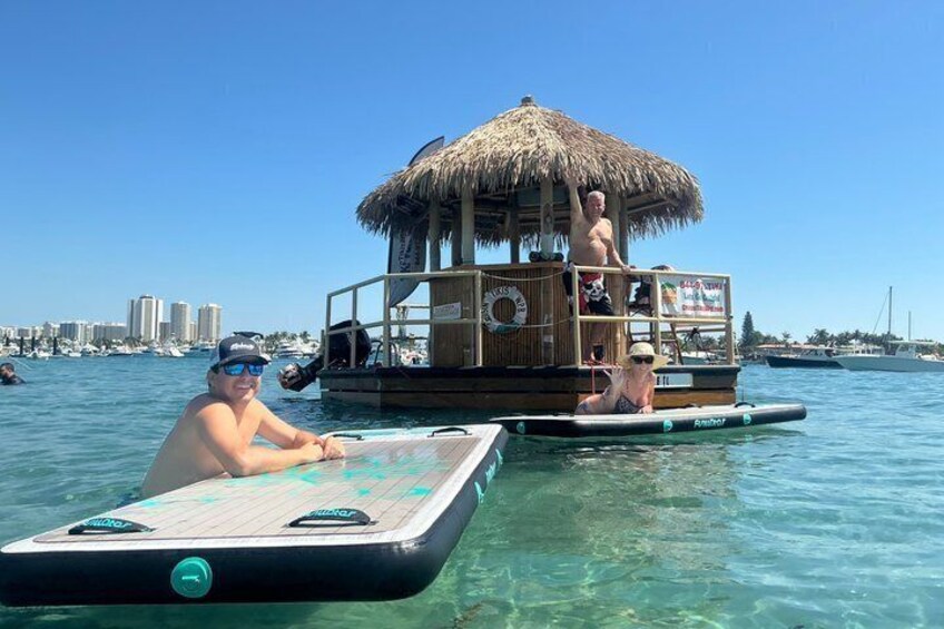 Un homme se tient sur une plateforme flottante surmontée d’un toit de chaume, un autre homme est sur une planche à pagaie, et on aperçoit la silhouette d’une ville en arrière-plan. 3