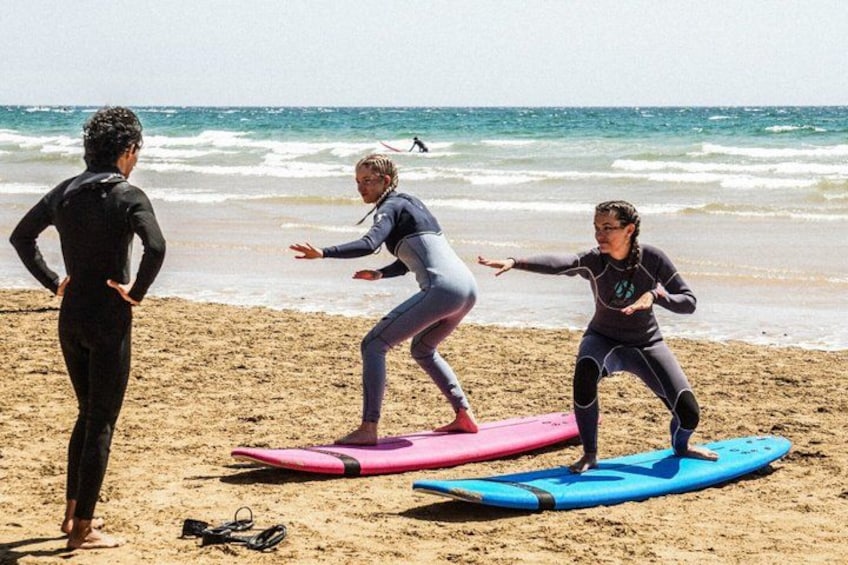 Tre personer på en strand med surfboards, hvoraf den ene står og to er på sandet. 4